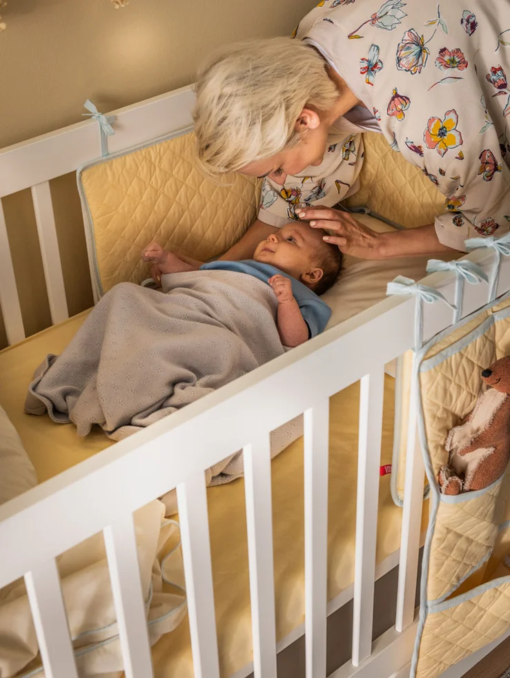Babcia pochyla się nad śpiącym niemowlakiem w białym łóżeczku z materacem.